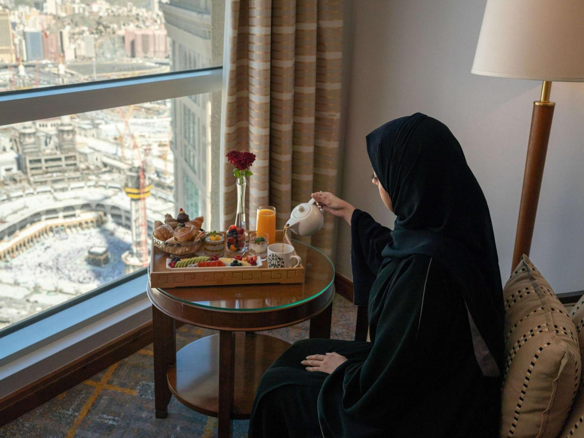 Swissotel Al Maqam Makkah Meca Exterior foto The image depicts a woman wearing a black abaya, sitting at a table near a large window. She is pouring tea from a teapot into a cup. The table has a beautifully arranged tray with various food items, including pastries and fruits, as well as a glass