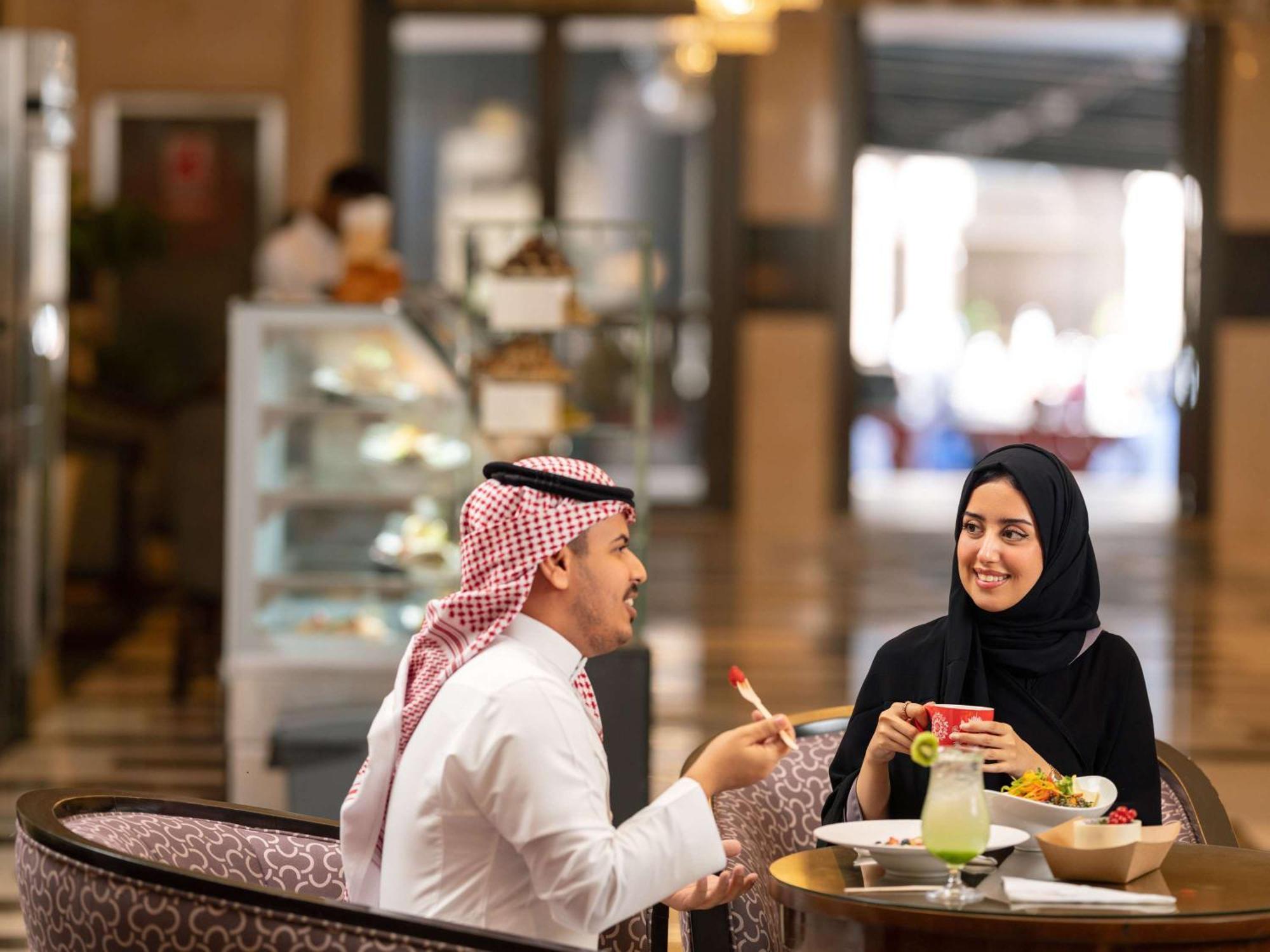 Swissotel Al Maqam Makkah Meca Exterior foto The photo features a man and a woman engaged in a conversation at a dining table. The man is wearing traditional attire with a white thobe and a headscarf, while the woman is dressed in a black abaya and headscarf. They appear to be enjoying a meal t