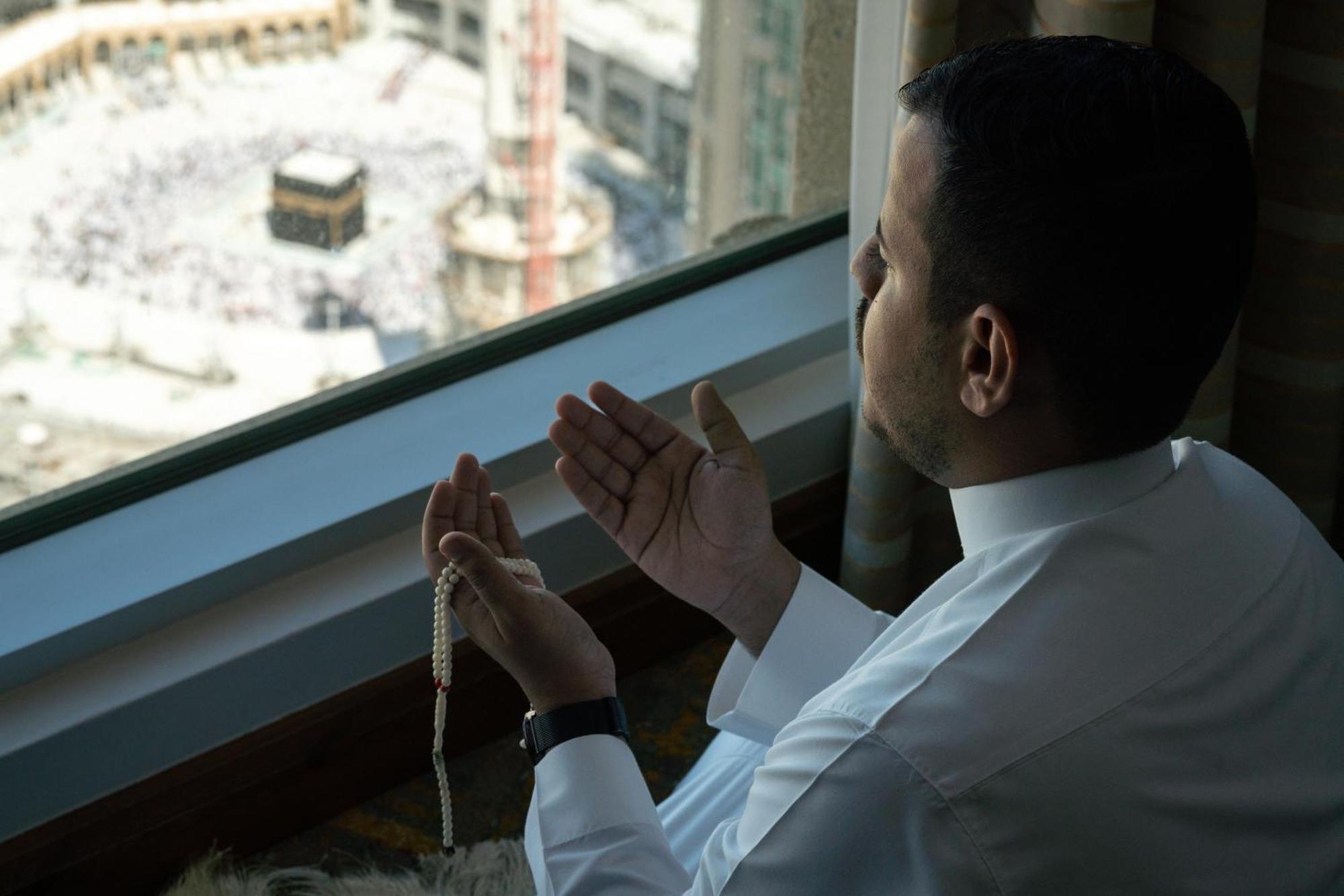 Swissotel Al Maqam Makkah Meca Exterior foto The photo shows a man sitting next to a window, engaged in prayer. He is facing toward the direction of the Kaaba, which is visible in the background, suggesting he is in Mecca. The man is holding prayer beads in one hand while gesturing in supplicat