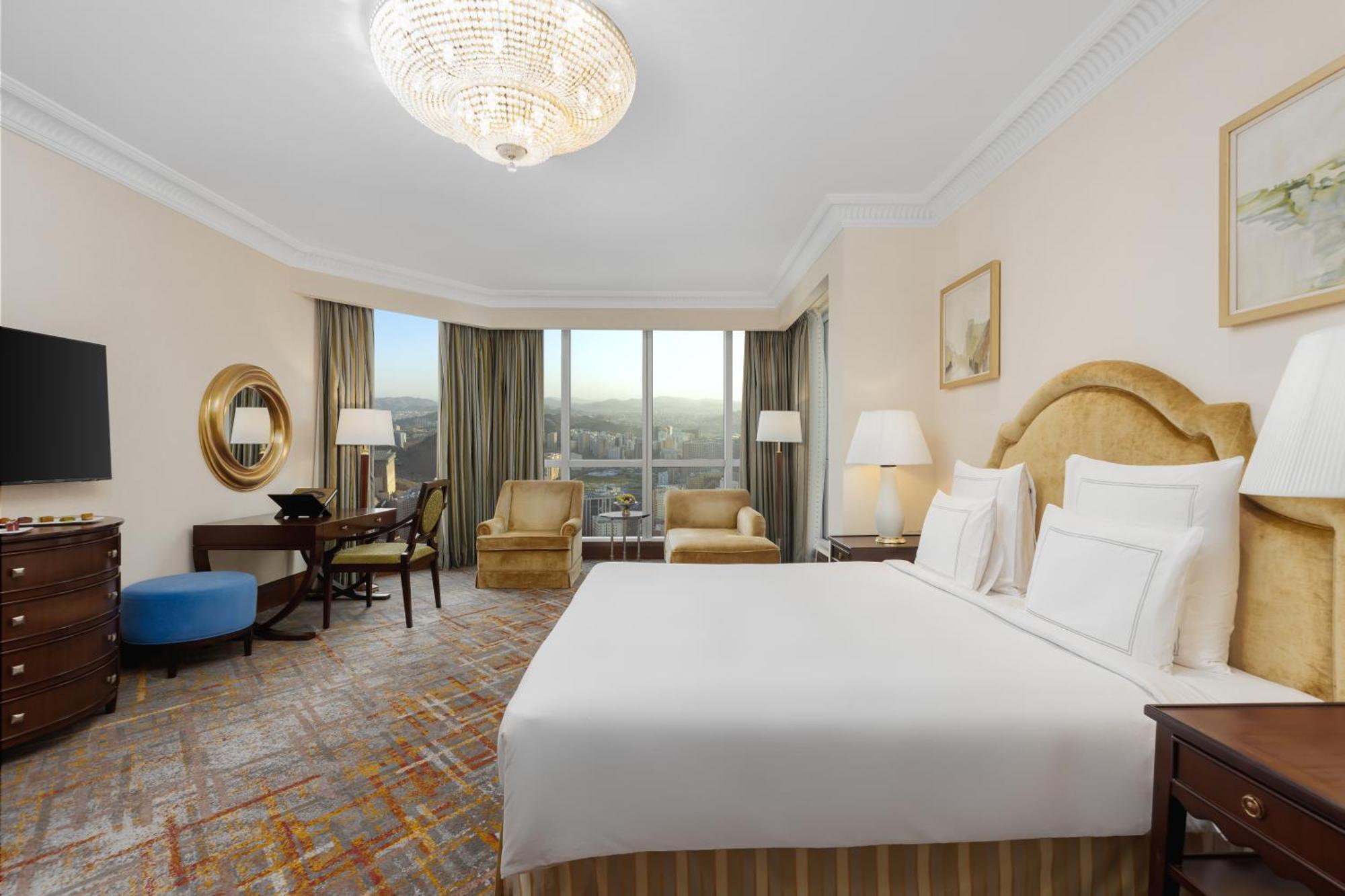 Swissotel Al Maqam Makkah Meca Exterior foto The photo shows a well-furnished hotel room with a modern and elegant design. It features a large bed with white linens and decorative pillows. There are two armchairs nearby, a small round table, and a writing desk with a chair. A television is moun