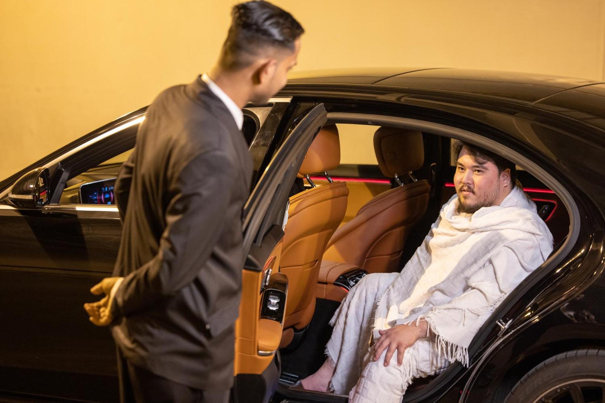 Swissotel Al Maqam Makkah Meca Exterior foto The image shows a scene outside a car at night. One person, dressed in a formal black suit, is standing next to an open car door, seemingly talking to the other person inside the car. The person inside the car appears to be wearing a white blanket or