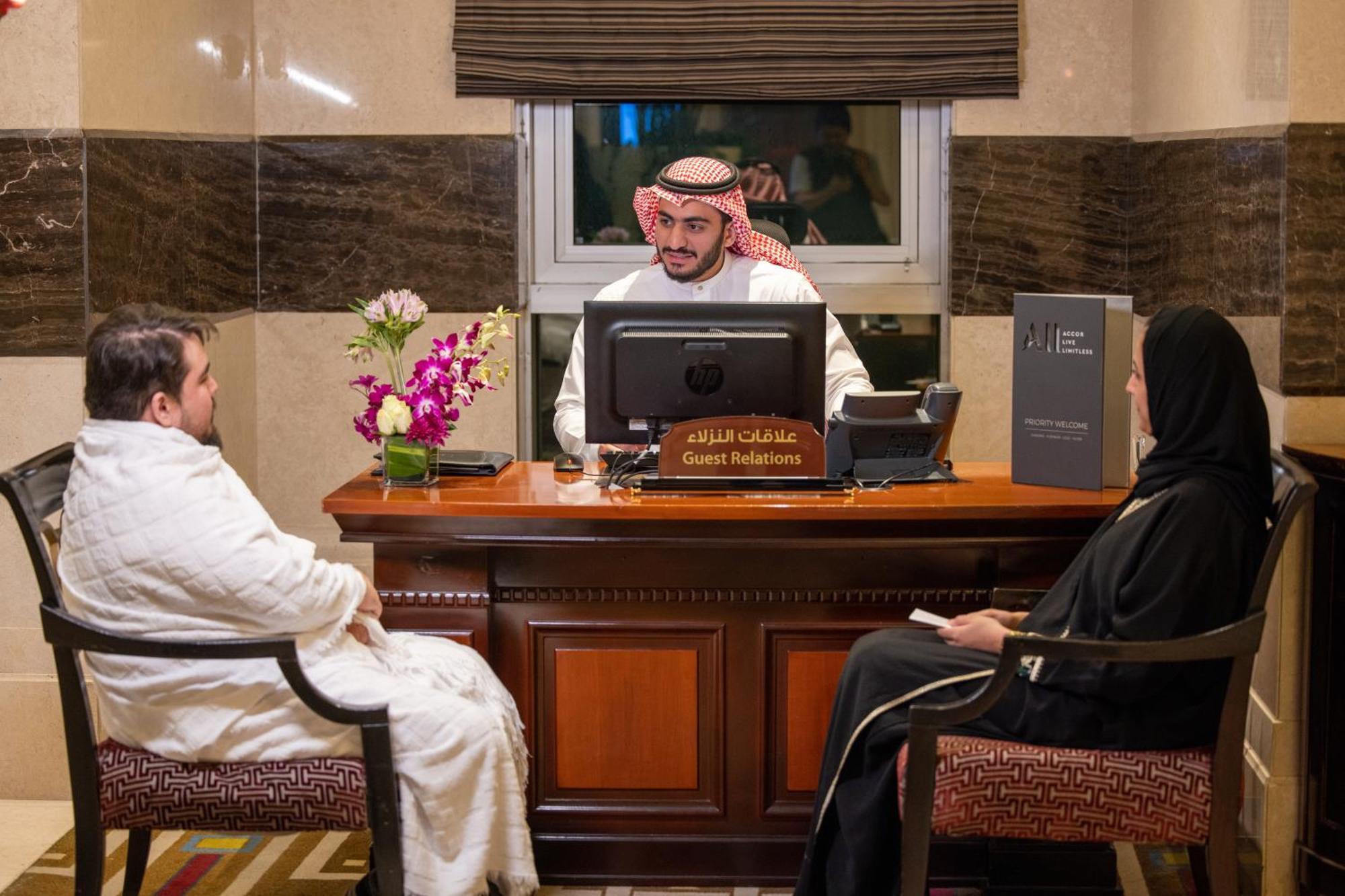 Swissotel Al Maqam Makkah Meca Exterior foto The photo shows a reception area with a male receptionist seated at a desk, wearing traditional attire, including a headscarf. He is focused on a computer screen. In front of him, there are two individuals seated: one is a man in traditional clothing