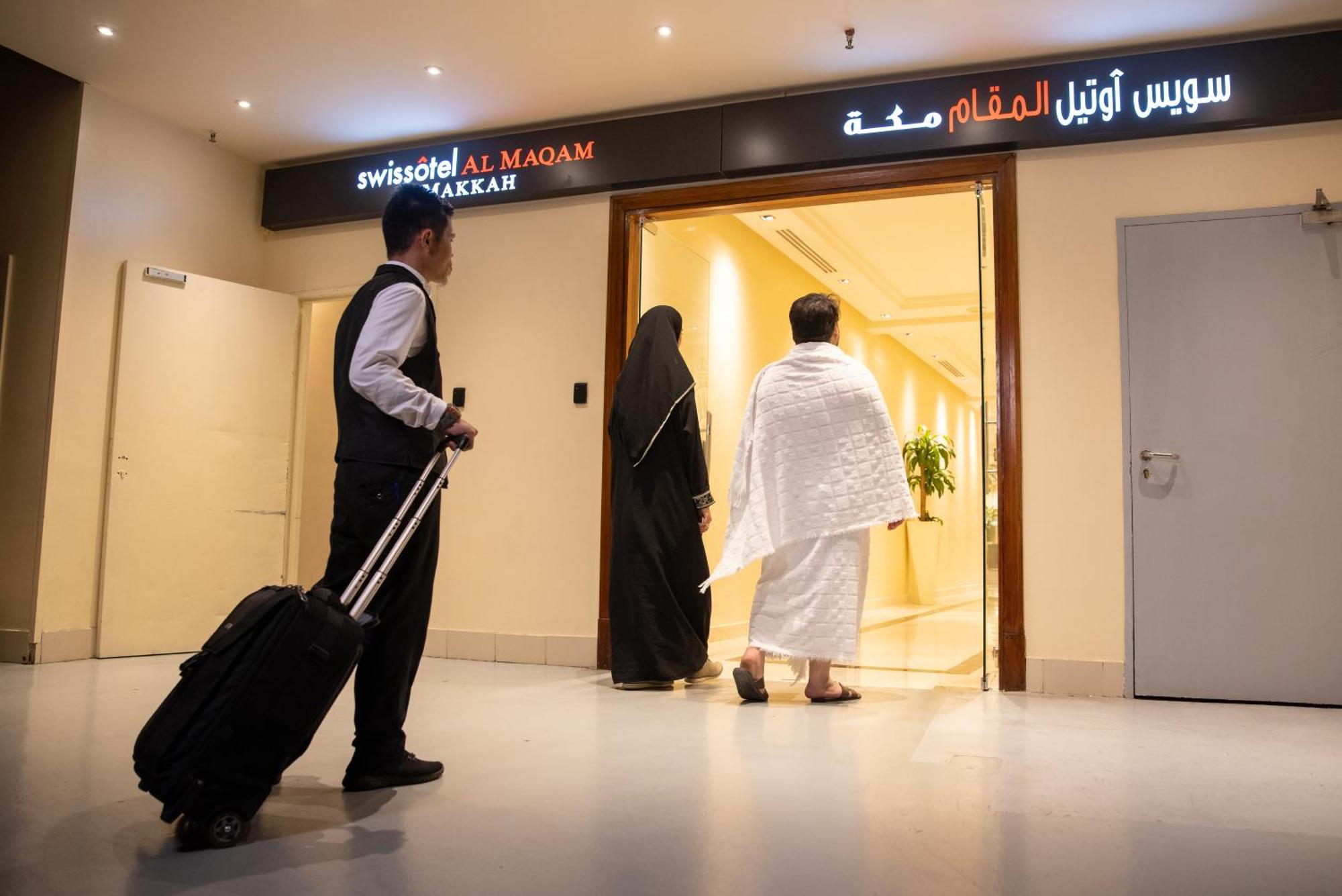Swissotel Al Maqam Makkah Meca Exterior foto The photo shows a hotel entrance, specifically for the "Swissotel Al Maqam Makkah," as indicated by the sign. In the scene, a man in a formal outfit carrying a suitcase waits nearby, while two individuals, one in traditional clothing (possibly a woma