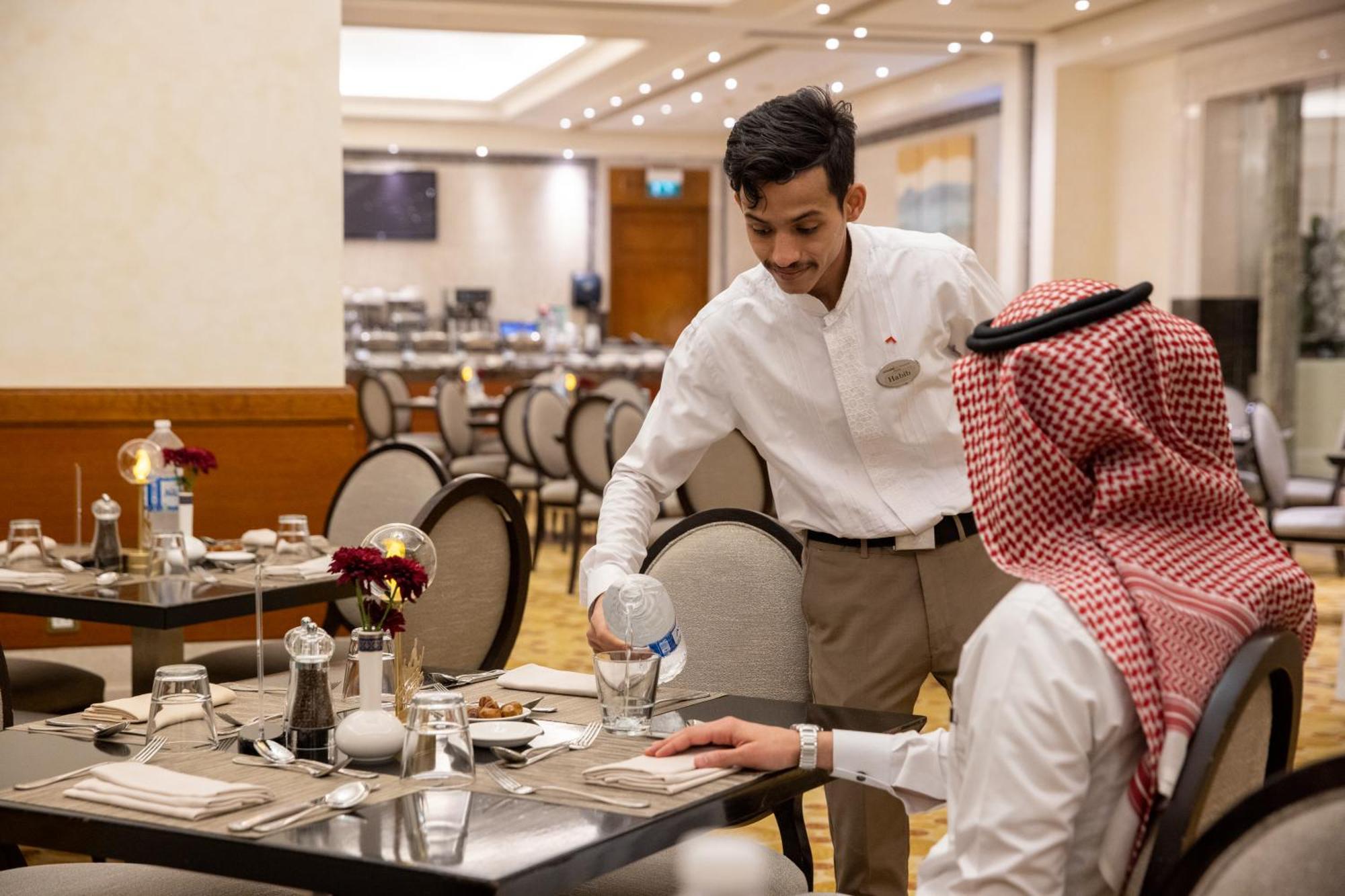 Swissotel Al Maqam Makkah Meca Exterior foto The photo shows a dining scene in a restaurant. A waiter, dressed in a white shirt and light-colored pants, is pouring water into a glass placed on the table. The table is set with plates, cutlery, and a small vase with flowers. The diner, who is wea