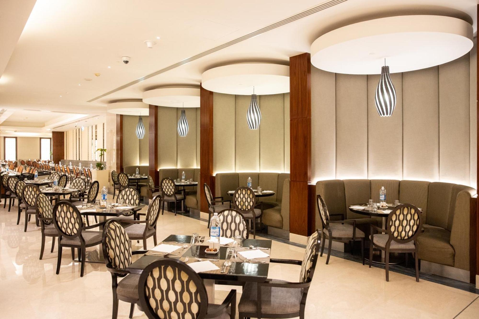 Swissotel Al Maqam Makkah Meca Exterior foto The photo shows a stylish restaurant interior. It features several dining tables arranged neatly with elegant chairs that have a patterned design. The walls are adorned with soft, muted colors and have wooden accents. Ambient lighting is provided by 