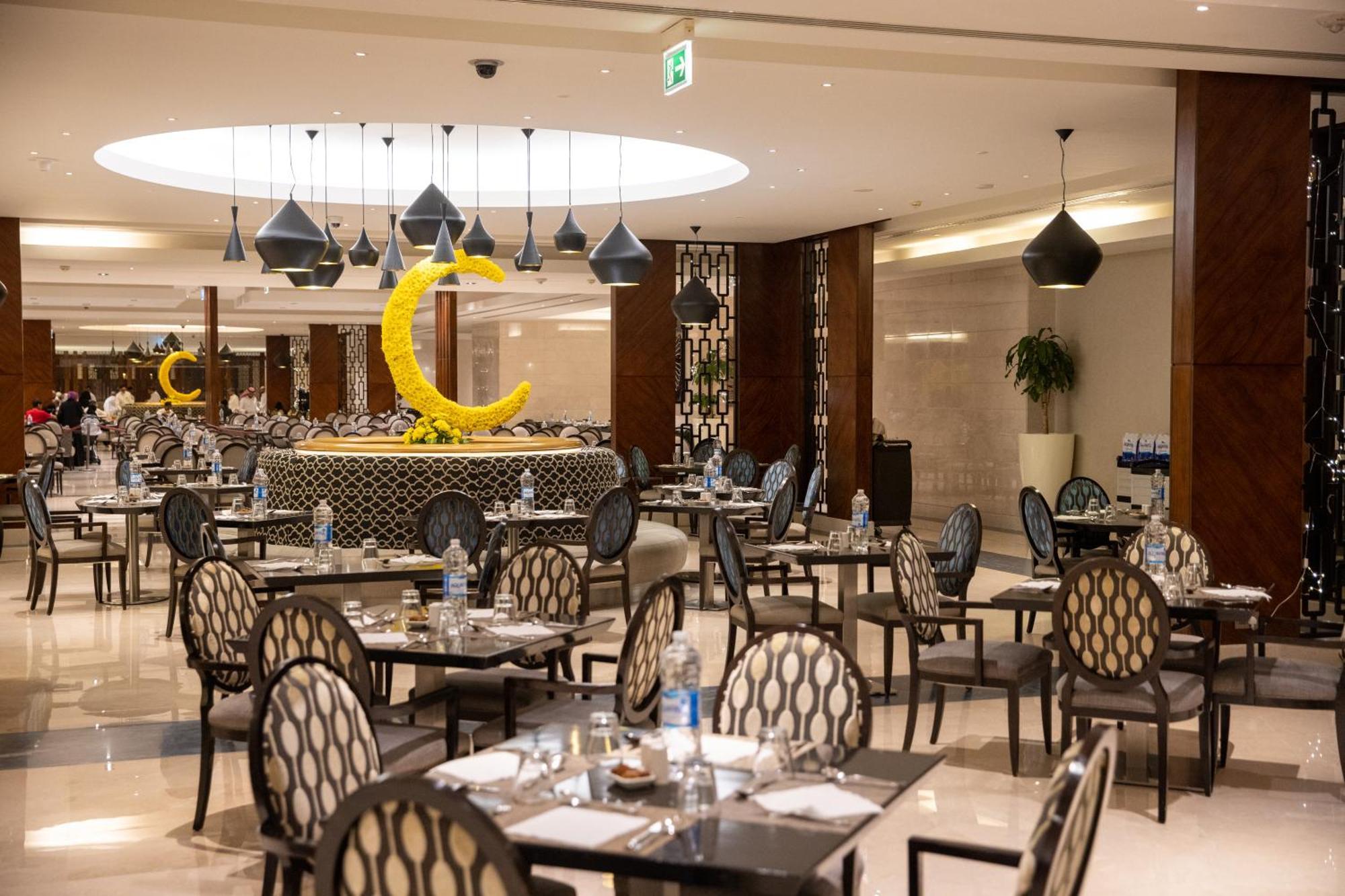 Swissotel Al Maqam Makkah Meca Exterior foto The photo shows the interior of a restaurant that features a modern and elegant design. The space has several tables arranged with stylish chairs that have a circular pattern. There are pendant lights hanging from the ceiling in various shapes, provi