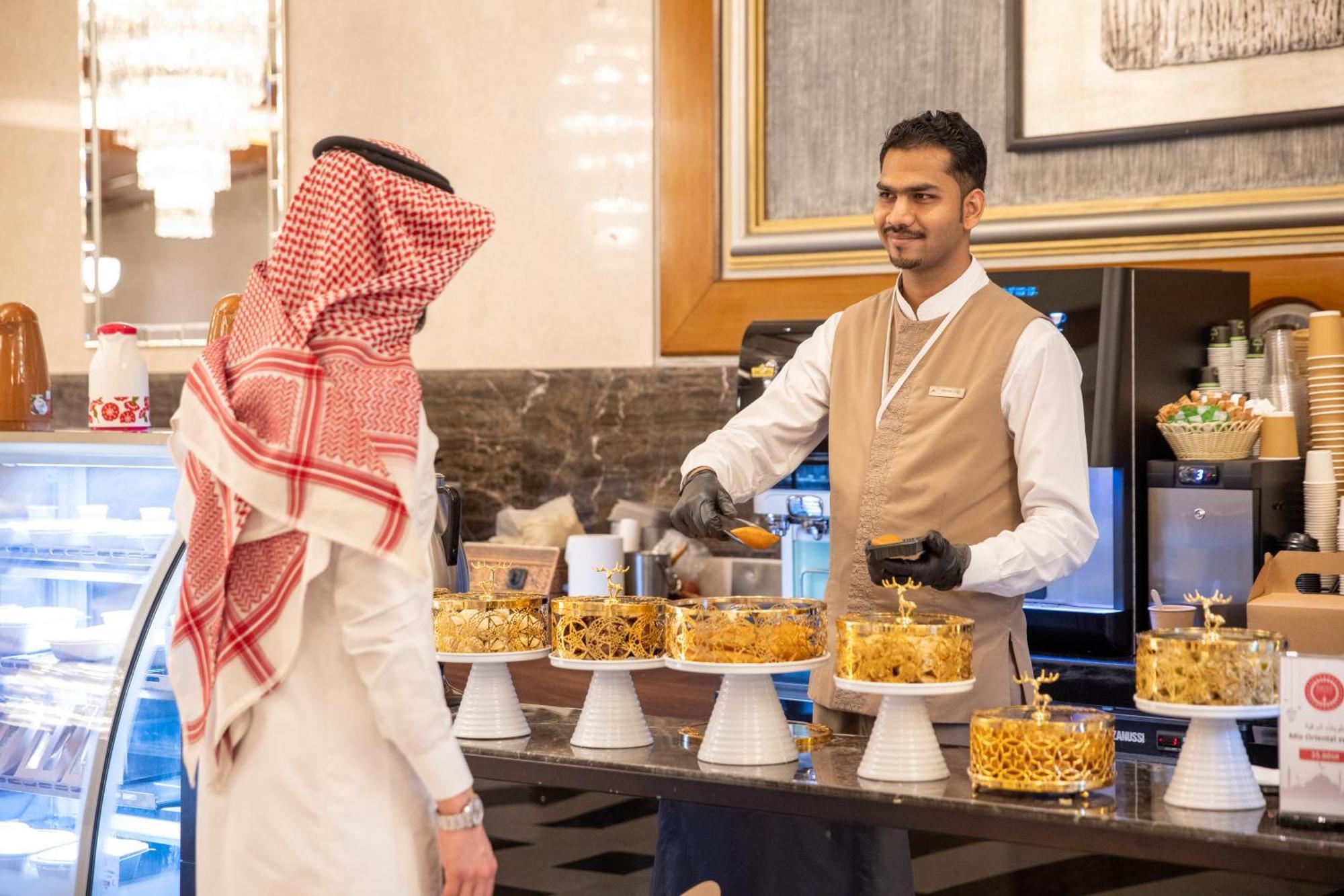 Swissotel Al Maqam Makkah Meca Exterior foto The photo shows a scene in a dessert display area, likely in a hotel or upscale restaurant. A man dressed in traditional Saudi attire (a white thobe and a red and white checkered headscarf) is facing a staff member who is wearing a beige uniform with