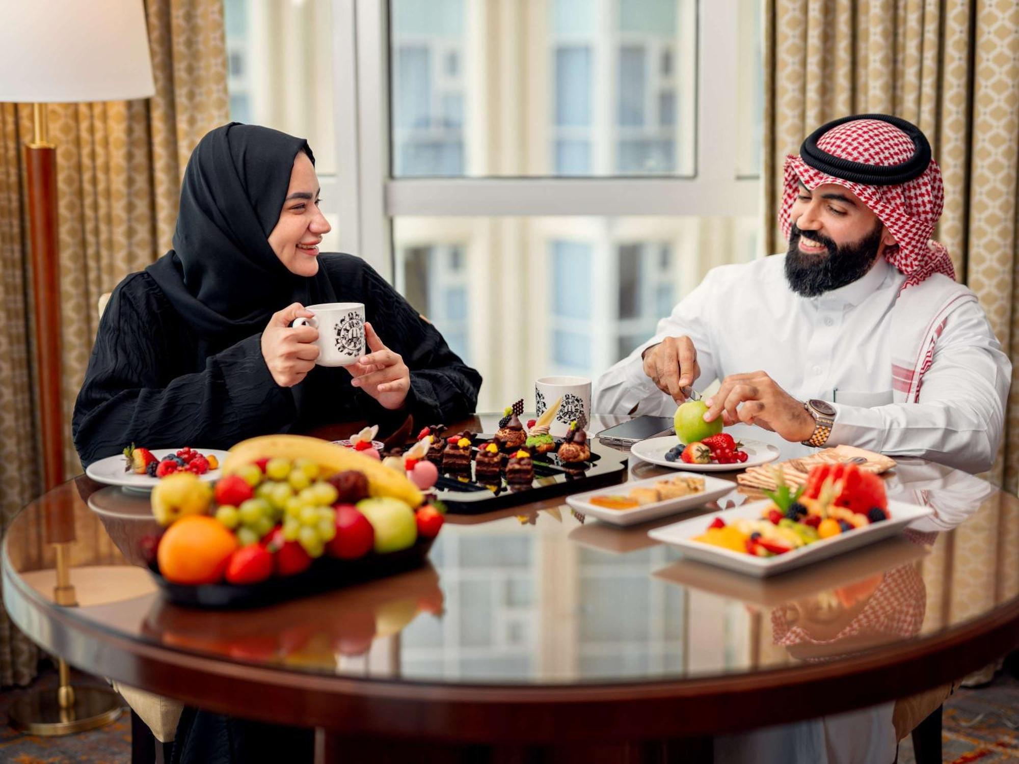 Swissotel Al Maqam Makkah Meca Exterior foto The photo shows two people enjoying a meal together at a dining table. The woman is wearing a black abaya and smiling while holding a cup, and the man is dressed in traditional white attire with a red and white checkered headscarf. They are surrounde