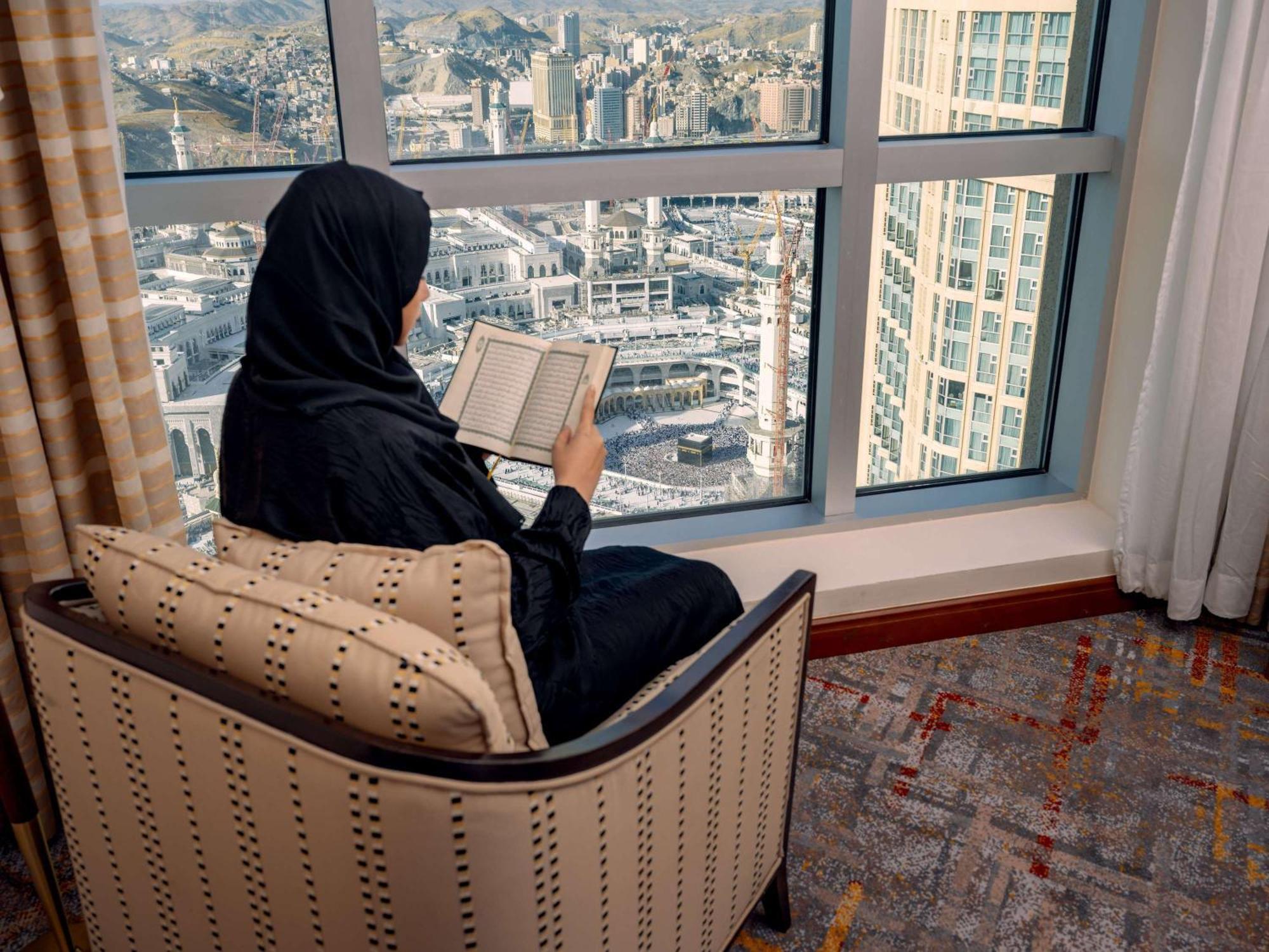 Swissotel Al Maqam Makkah Meca Exterior foto The photo depicts a woman seated in a comfortable chair by a large window. She is wearing a traditional black garment and is focused on reading a book. Outside the window, there is a view of a cityscape, possibly a metropolitan area, with various bui