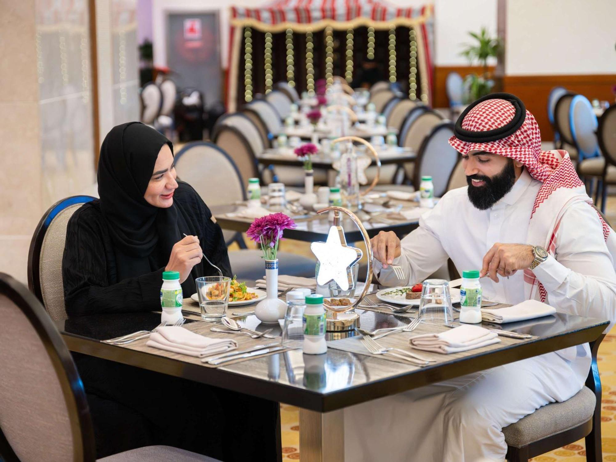Swissotel Al Maqam Makkah Meca Exterior foto The photo shows a couple dining at a restaurant. The man is wearing traditional attire, including a white thobe and a red and white checkered headscarf, while the woman is dressed in a black abaya and a headscarf. They are engaged in a pleasant conve