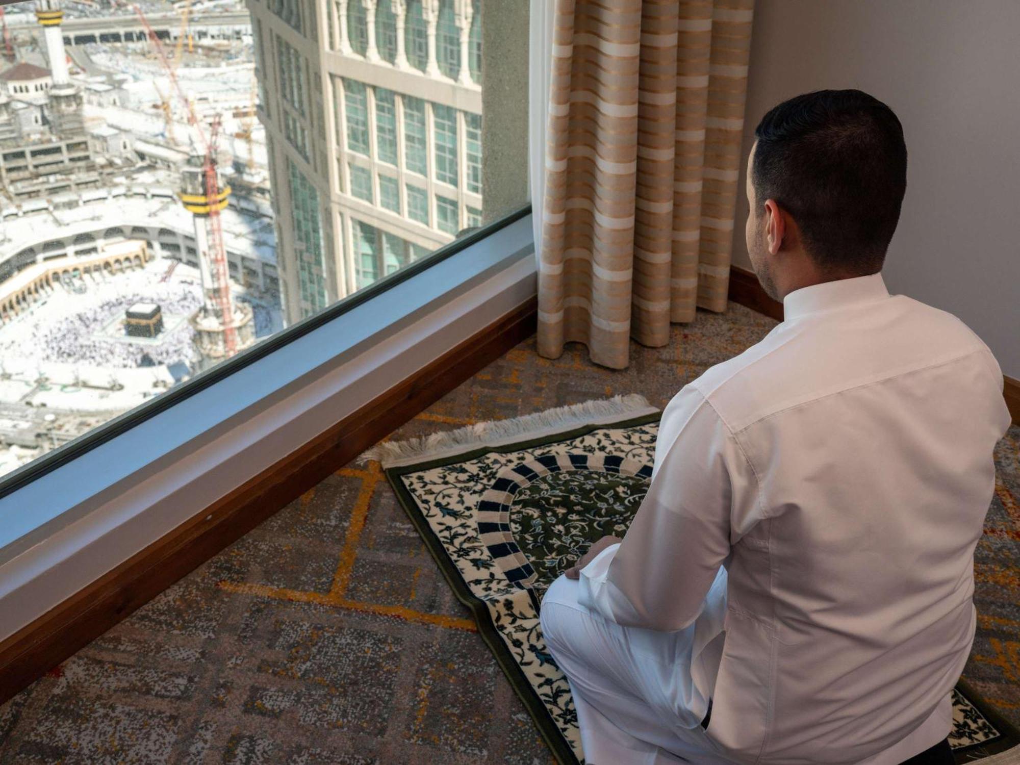 Swissotel Al Maqam Makkah Meca Exterior foto The photo shows a man sitting in a prayer position on a prayer mat, facing a large window. Through the window, there's a view of a construction site, possibly near a significant religious site, given the context. The man is wearing traditional attire