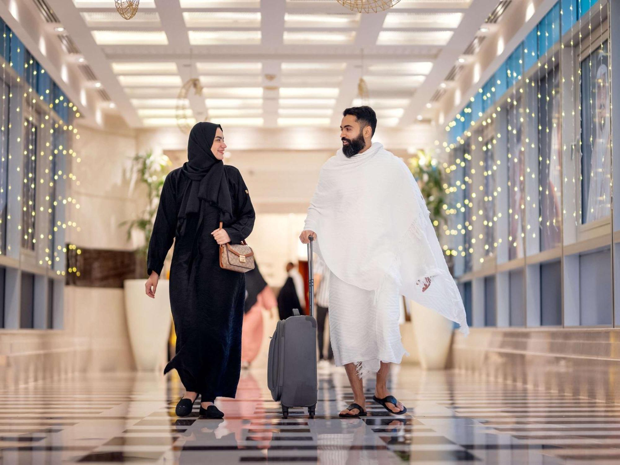 Swissotel Al Maqam Makkah Meca Exterior foto The photo depicts a couple walking together in a modern, well-lit hallway. The man is dressed in traditional white garments, while the woman is wearing a black abaya. The hallway features a stylish marble floor and is adorned with decorative lights, 