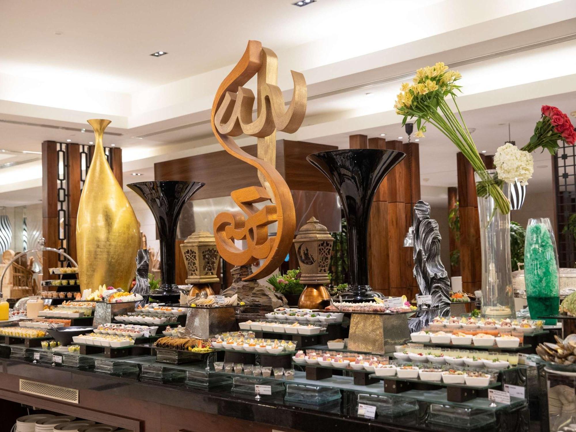 Swissotel Al Maqam Makkah Meca Exterior foto The photo shows a beautifully arranged buffet display in a restaurant. In the center, there is a large decorative piece featuring Arabic calligraphy. Surrounding it are various elegant food presentations, including trays of desserts, likely reflectin