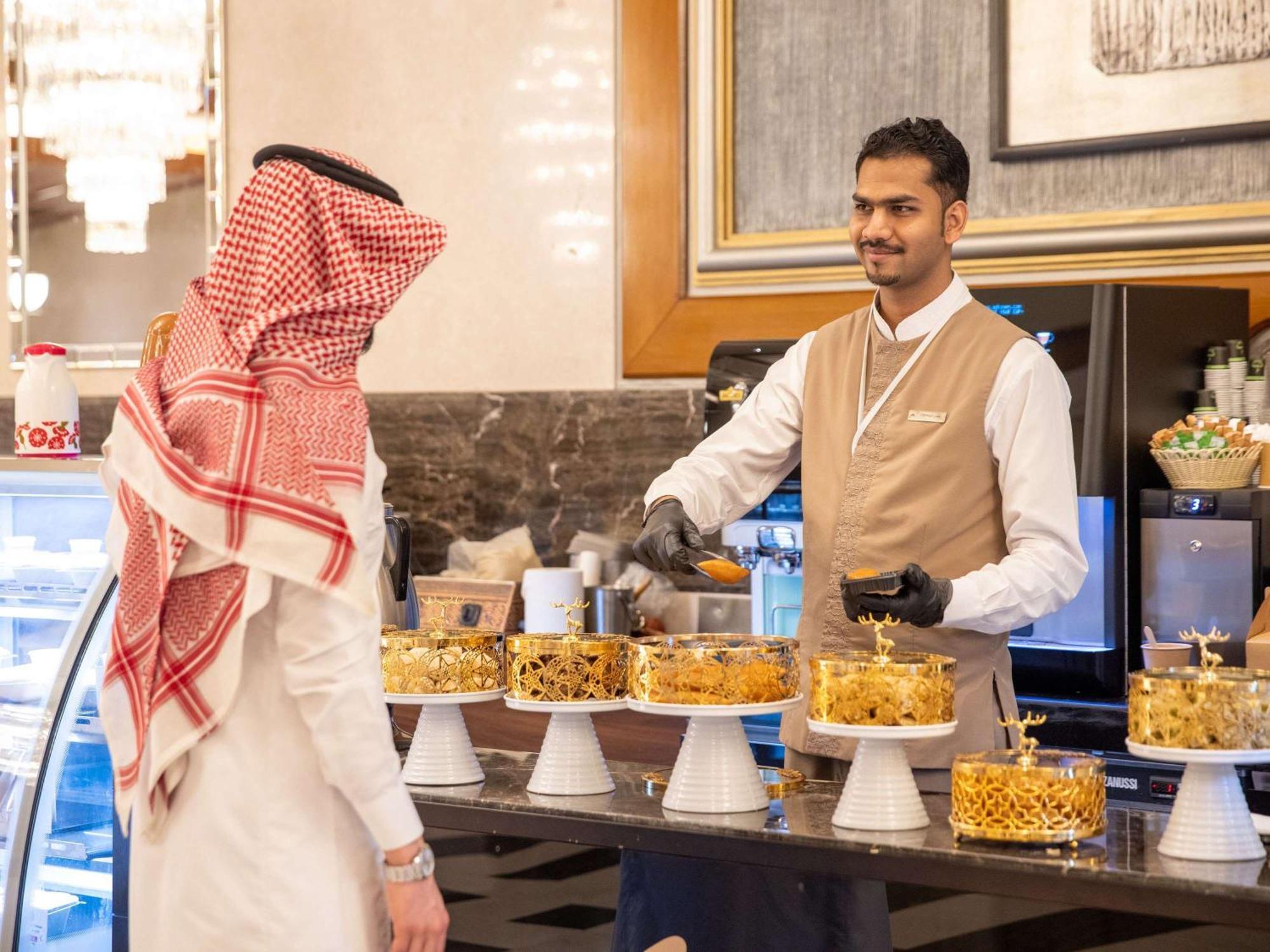 Swissotel Al Maqam Makkah Meca Exterior foto The photo depicts a scene in a bakery or dessert shop. There is a display of various beautifully decorated cakes arranged on stands. A man wearing traditional attire, possibly a thobe and a headscarf, is interacting with a staff member who is smiling